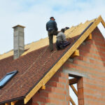 Two roofers are installing an asphalt shingle roof on a newly constructed brick home.